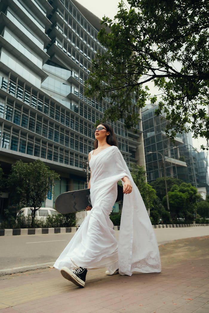 A woman in a white saree and sneakers holds a skateboard, walking through a modern urban environment.